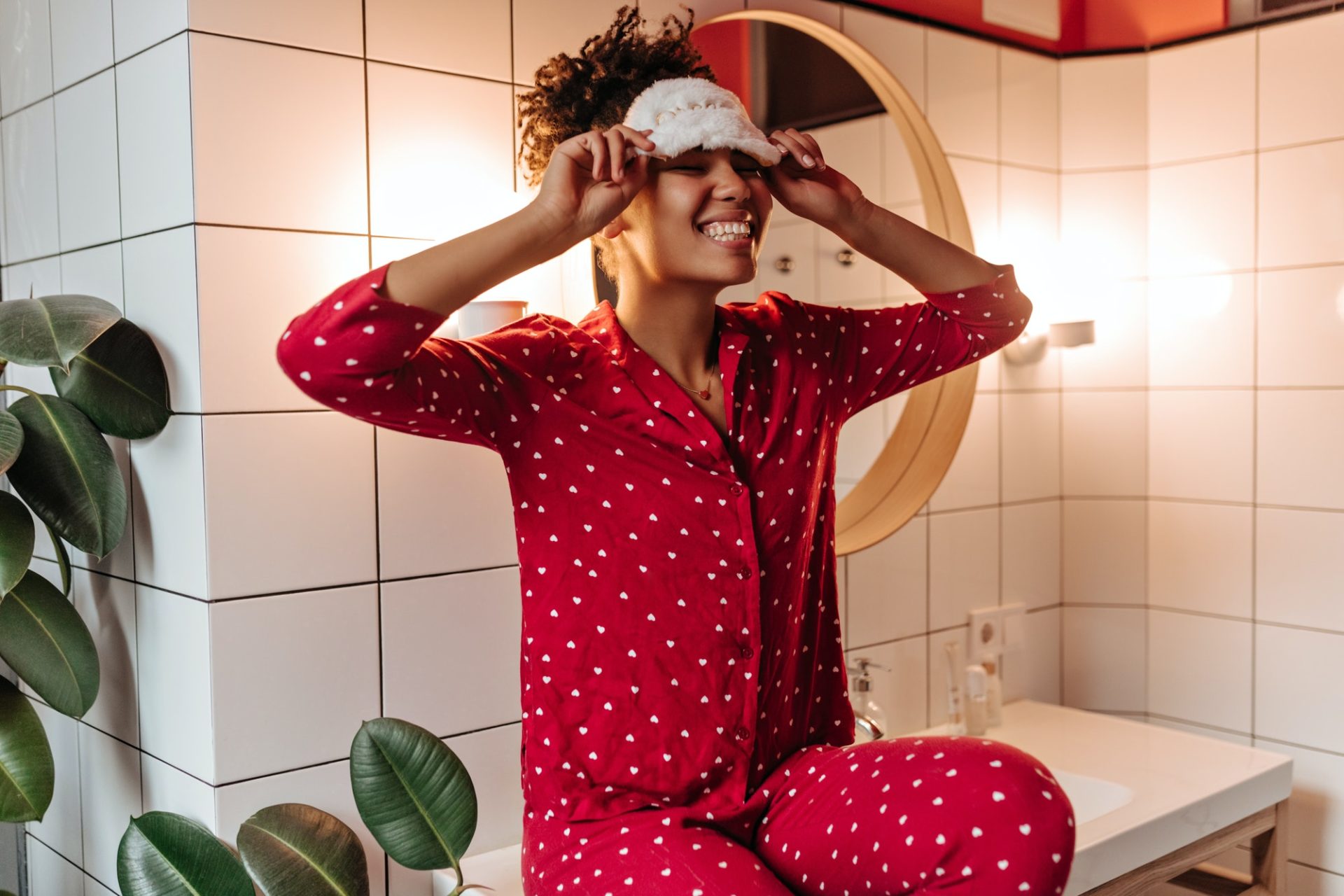 Emotional woman in pajamas laughs, takes off her sleep mask and sits near bathroom mirror