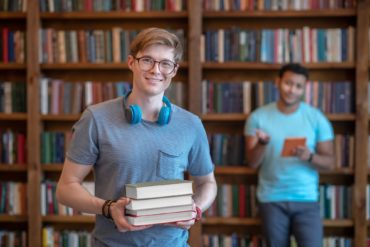 Fair-haired young man in eyeglasses standing in the library with his friend
