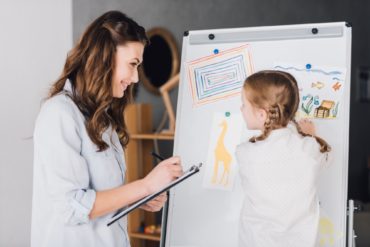 happy psychologist with clipboard talking to child near whiteboard with various drawings