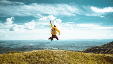Hiker with backpack raising hands jumping on the top of a mountain