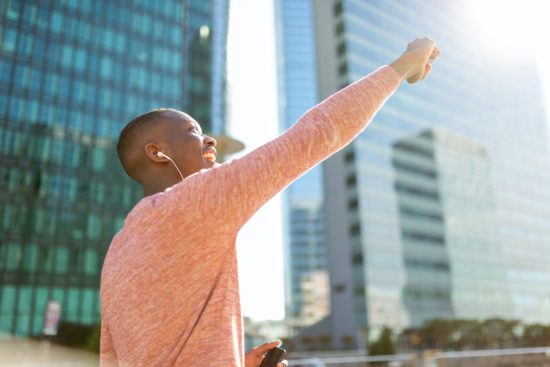 Side of young black man punching the air, listening to music with earphones and cellphone